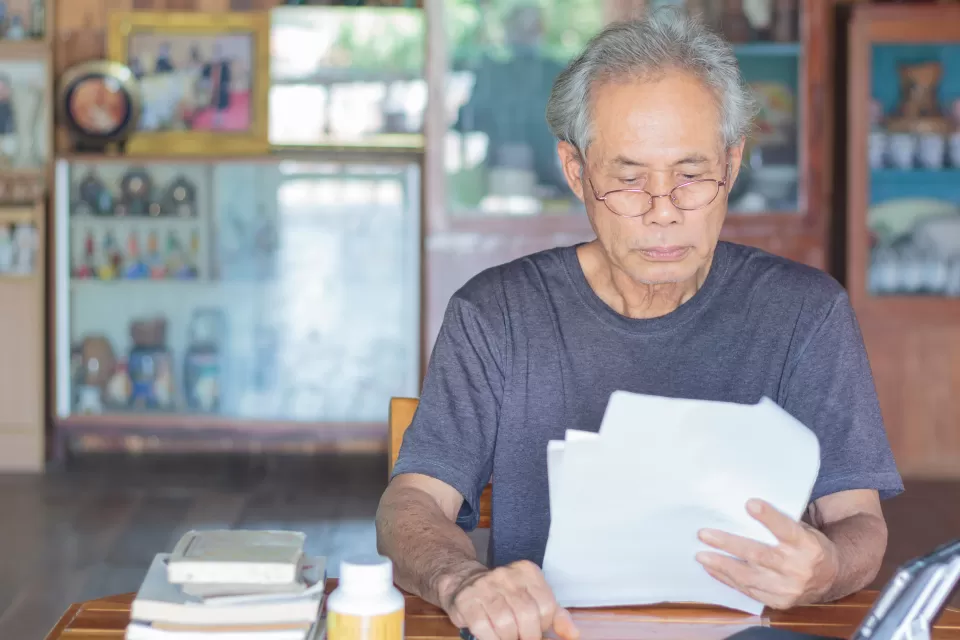 Gray-haired Asian man wearing glasses sitting at wooden table reading documents